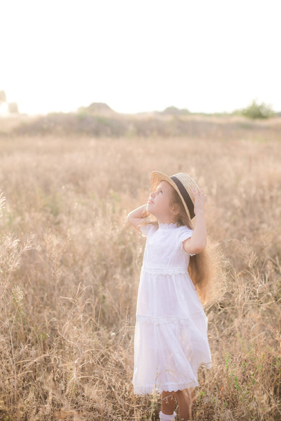 Słodka mała dziewczynka z długimi blond kręconymi włosami w białej letniej sukience i słomkowym kapeluszem na polu na wsi w lecie o zachodzie słońca. Natura i ekożycie - Zdjęcie, obraz