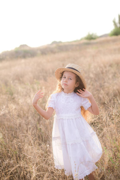 Милая маленькая девочка с длинными светлыми вьющимися волосами в белом летнем платье и соломенной шляпкой на поле в сельской местности летом на закате. Природа и экожизнь - Фото, изображение