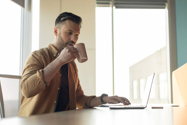 Erfolgreicher Geschäftsmann arbeitet online am Laptop und trinkt Kaffee im Büro - Foto, Bild