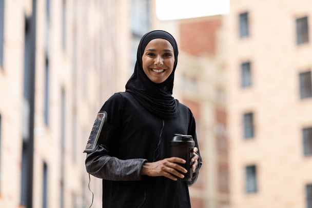Счастливая молодая арабская женщина в хиджабе и спортивной форме с телефоном на плече с чашкой коктейля - Фото, изображение