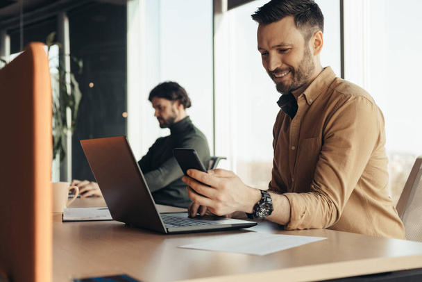 Glückliche männliche Firmenangestellte checkt Smartphone, nutzt Laptop, arbeitet mit männlichen Kollegen im modernen Büro - Foto, Bild