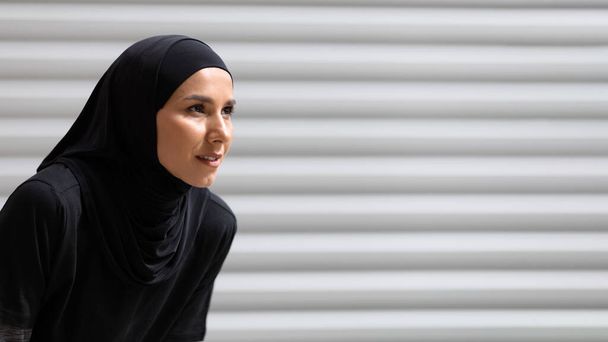Müde ernsthafte hübsche junge arabische Dame im Hijab, die sich von Ausdauerübungen auf grauem Wandhintergrund ausruht, Nahaufnahme - Foto, Bild