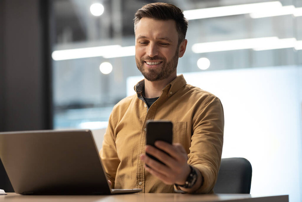 Ευτυχισμένος επιχειρηματίας κρατώντας το κινητό μήνυμα εργασίας σε φορητό υπολογιστή στο γραφείο - Φωτογραφία, εικόνα