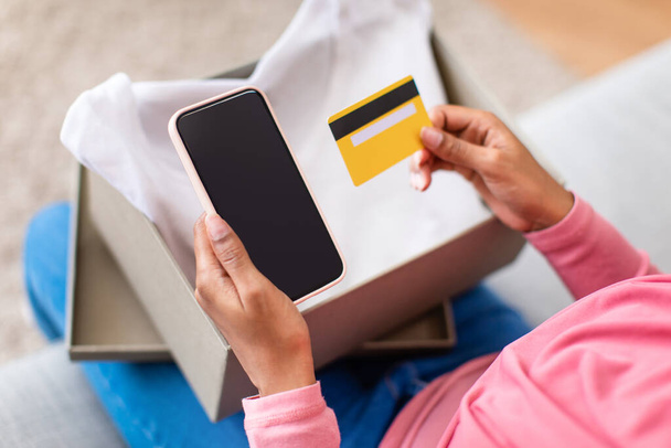 Mujer afroamericana con caja de cartón, mostrando teléfono inteligente con pantalla en blanco y tarjeta de crédito, maqueta - Foto, Imagen