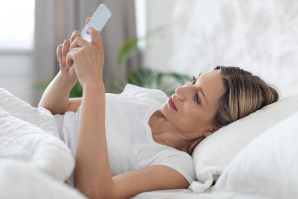 Señora positiva acostada en la cama sola, usando teléfono móvil - Foto, Imagen