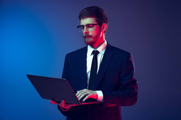 Portrait of young stylish serious man in business suit working remote isolated on dark blue studio background. Concept of emotions, work, sales, ad, fashion and beauty - 写真・画像