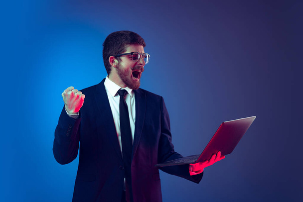 Удивительный молодой человек в деловом костюме и очках, держащий ноутбук и кричащий изолированно на темно-синем фоне. Эмоции, работа, продажи, реклама, ставки - Фото, изображение