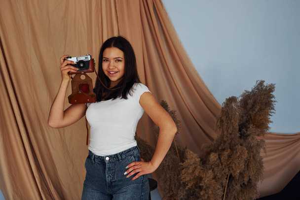 Young cheerful woman holds indoors and holds retro camera. - Фото, изображение