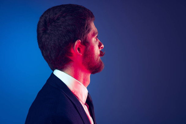 Closeup. Profile view of young stylish bearded man in business suit posing isolated on dark blue studio background. Concept of emotions, style, sales, ad, fashion and beauty - Valokuva, kuva