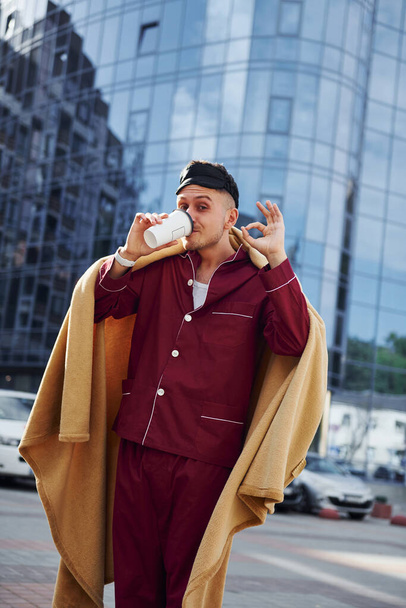 Morning coffee cup. Young man in pajama is outdoors on the street. Feels sleepy. - Fotó, kép