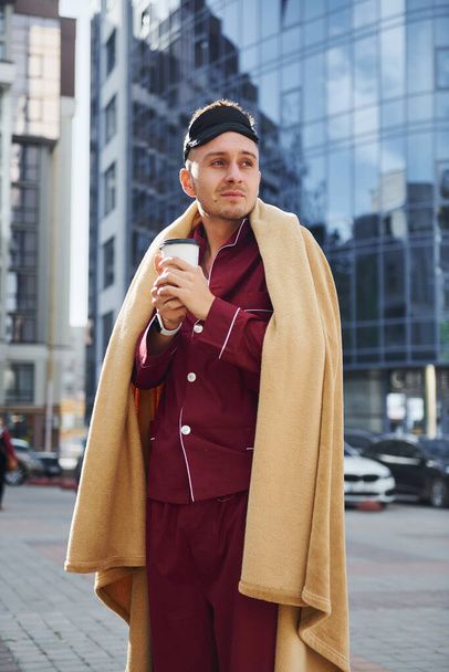 Morning coffee cup. Young man in pajama is outdoors on the street. Feels sleepy. - Fotó, kép
