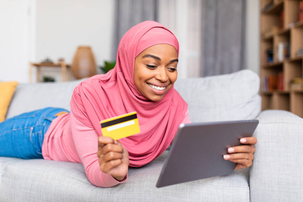 Mujer musulmana afroamericana feliz acostada en el sofá y haciendo compras en línea, usando tableta digital y tarjeta de crédito - Foto, Imagen