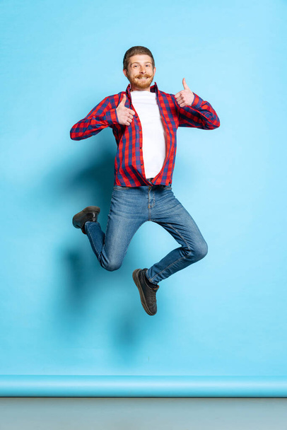 Emotional young red-headed man in white t-shirt and plaid shirt jumping isolated on blue background. Concept of art, fashion, emotions, aspiration - Foto, afbeelding