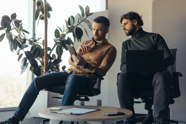 Colaboradores masculinos enfocados que cooperan, discutiendo ideas de negocios con computadora portátil y tableta digital en la oficina de la compañía - Foto, Imagen