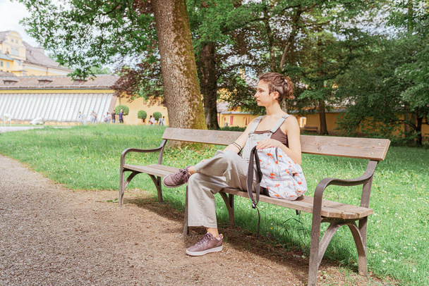 Istun puistonpenkillä. nuori kaunis teini tyttö, hiukset sidottu pulla, jossa oyukzak, istuu puistossa penkillä. Kaunis tyttö ulkona kesäpäivänä. luonto tausta. - Valokuva, kuva