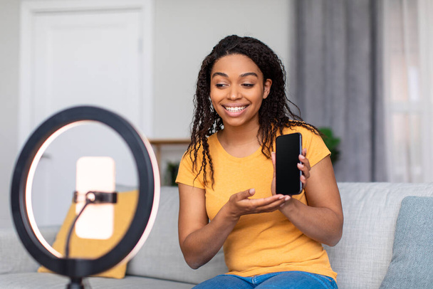 アフリカ系アメリカ人女性のインフルエンサーが自宅で携帯電話のウェブカメラで現代のスマートフォンのビデオレビューを撮影 - 写真・画像