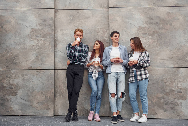 Group of young positive friends in casual clothes standing together against grey wall with cups of drink in hands. - 写真・画像