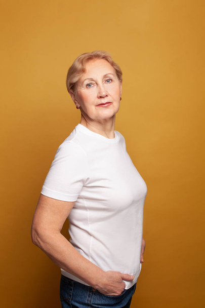 Blonde model senior woman in white t-shirt standing on bright yellow studio wall background - Φωτογραφία, εικόνα
