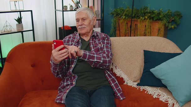 Senior old grandparent works on mobile phone, sends messages, makes online purchases at home sofa - Фото, изображение