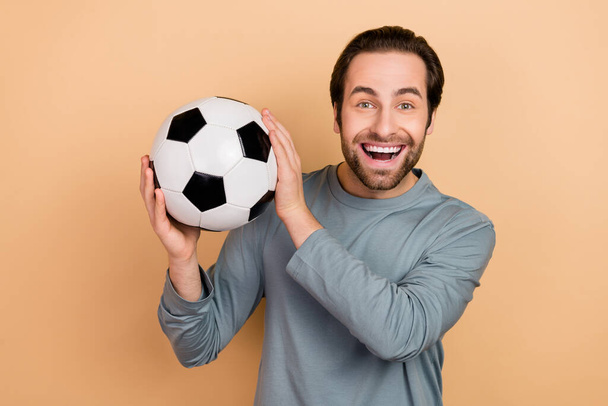Portrait of satisfied glad man arms hold football toothy smile look camera isolated on beige color background - 写真・画像
