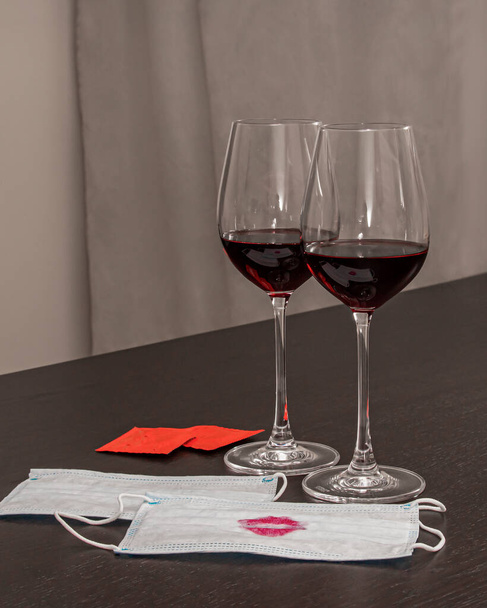 δύο ποτήρια κρασί, δύο προστατευτικές μάσκες και δύο προφυλακτικά είναι στο τραπέζι ως σύμβολο μιας ρομαντικής βραδιάς για τους εραστές - Φωτογραφία, εικόνα