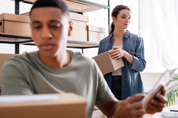 Young seller holding carton box near blurred african american colleague with digital tablet in online web store  - Foto, Imagem