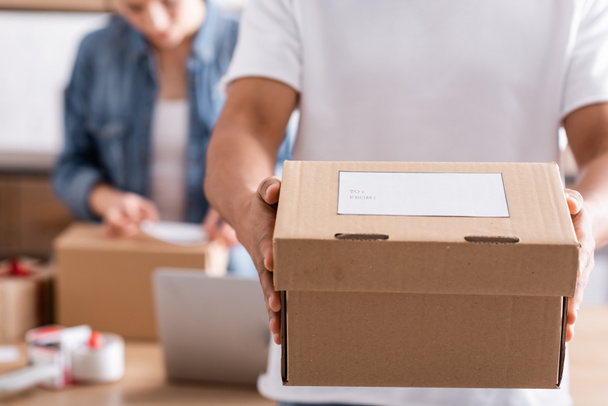 Cropped view of african american seller holding box with shipping label near blurred colleague in online web store  - Photo, Image