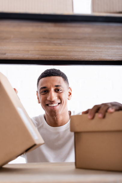 Gülümseyen Afrikalı Amerikalı satıcı internetteki raftaki bulanık karton kutuların yanındaki kameraya bakıyor.  - Fotoğraf, Görsel