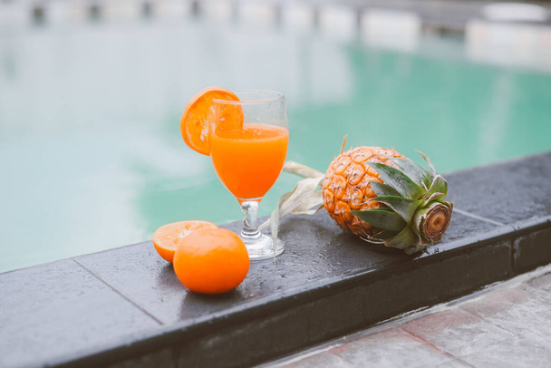 Bicchiere di succo sul bordo piscina, succo d'arancia con frutta all'arancia e ananas in piscina - Foto, immagini