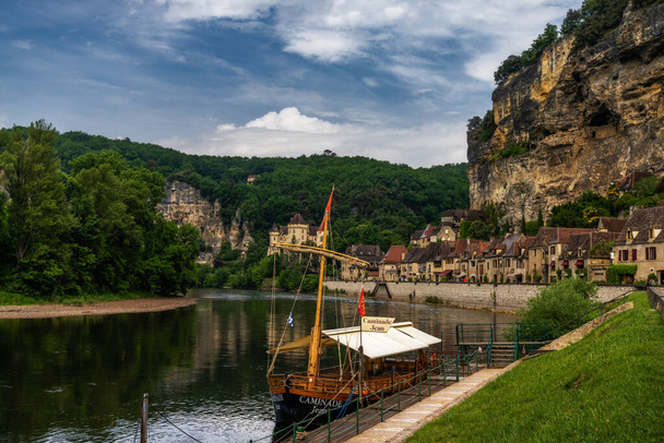 La Roque-Gageac, Francia - 12 maggio, 2022: veduta dello storico villaggio di La Roque-Gageac con il fiume Dordogna e le case a strapiombo e una barca di legno del Gabare in primo piano - Foto, immagini