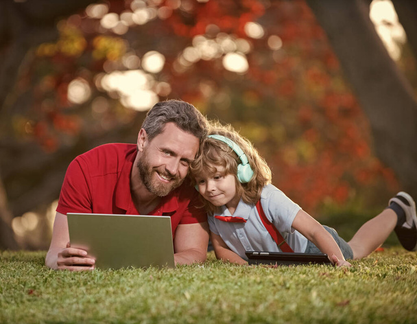 happy family of dad and son use laptop for lesson listen music in headphones in park, childhood. - Фото, изображение