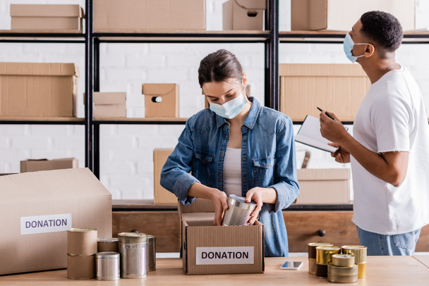 Multiethnic sellers in medical masks putting canned food in box with donation lettering in online web store  - Foto, afbeelding