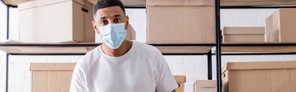 vendedor afroamericano en máscara médica mirando la cámara cerca de cajas en rack en la tienda web en línea, pancarta  - Foto, imagen