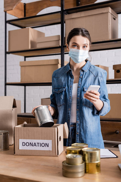 Seller in medical mask holding smartphone and canned food near donation box in online web store  - Photo, Image
