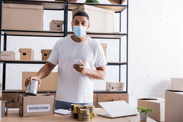 Venditore afroamericano in maschera medica con smartphone vicino scatola di cibo in scatola e donazione nel negozio online  - Foto, immagini