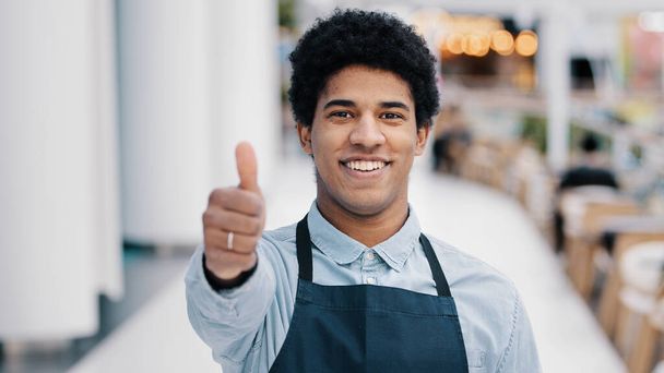 Φιλικός χαρούμενος Αφρικανός Αμερικανός εργαζόμενος σερβιτόρος πωλητής σε ποδιά δείχνει καλό σημάδι με το χέρι μικρή επιχείρηση ιδιοκτήτης του εστιατορίου καφέ δείχνει σαν αντίχειρας κοιτάζοντας κάμερα - Φωτογραφία, εικόνα