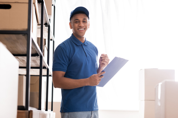 Positive african american deliveryman holding clipboard near rack with boxes in online web store  - Foto, Bild