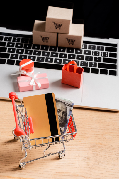Credit card and money in toy shopping cart near laptop with small packages on table  - Photo, Image