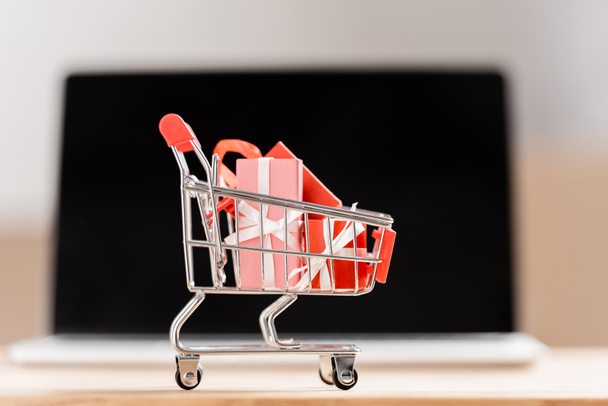 Toy shopping cart with gift boxes near blurred laptop  - Фото, зображення