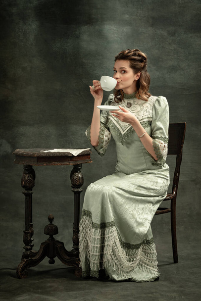 Vintage portrait of young beautiful girl in gray dress of medieval style drinking tea isolated on dark background. Comparison of eras concept, flemish style. Art, beauty - Fotografie, Obrázek