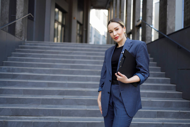 Elegance businesswoman with a clipboard folder in her hands stands on the stairs of a modern building - Foto, afbeelding