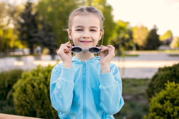 Una linda niña con una chaqueta azul y gafas de sol se sienta en un banco del parque y sonríe. Niño feliz de moda con coletas poses lindas para el fotógrafo al atardecer. Espacio libre para la prueba - Foto, Imagen