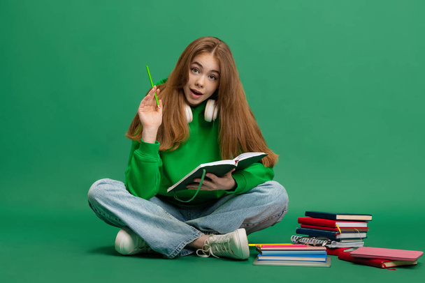 Portrait of young girl, student in casual cloth, hoodie sitting on floor, studying isolated over green studio background. Making notes - Fotó, kép