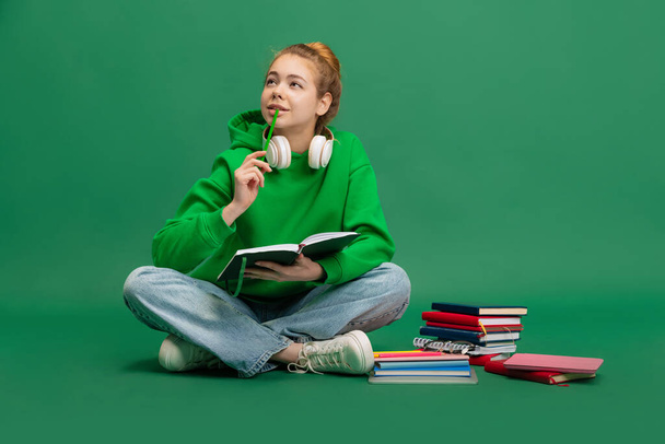 Portrét mladé dívky, student v ležérní látky sedí na podlaze se zamyšleným výrazem, studuje izolované přes zelené pozadí studia - Fotografie, Obrázek