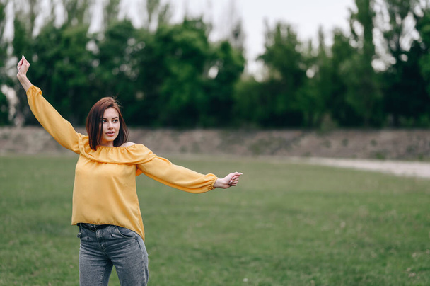 Ein Mädchen in gelber Bluse breitet die Arme zur Seite aus und blickt in die Ferne, inmitten einer grünen Wiese stehend.. - Foto, Bild