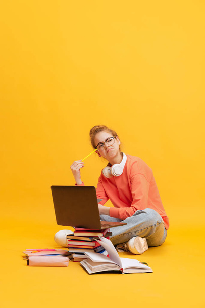 Retrato de menina, estudante de camisola e óculos sentados no chão com expressão pensativa, estudando isolado sobre fundo amarelo - Foto, Imagem