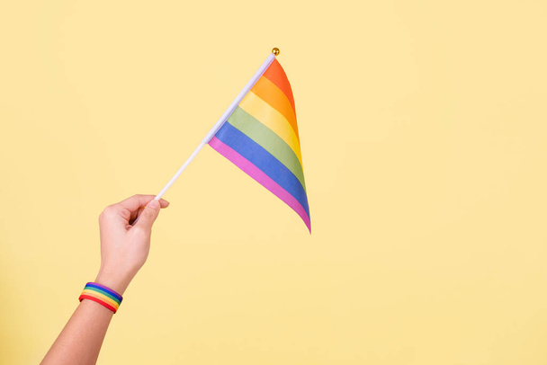Primer plano de una mano ondeando una pequeña bandera del arco iris contra aislado sobre un fondo amarillo - Foto, imagen