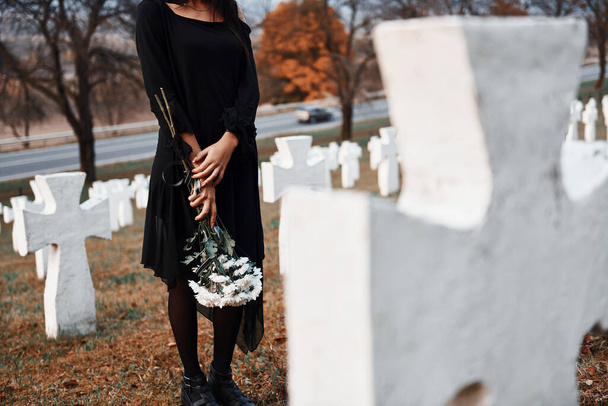Con fiori in mano. Giovane donna in abiti neri visita cimitero con molte croci bianche. Concezione di funerale e morte. - Foto, immagini