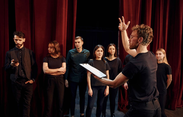 Man with notepad practice his role. Group of actors in dark colored clothes on rehearsal in the theater. - Foto, Imagen