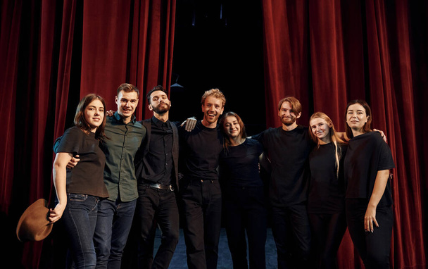 Standing against red curtains. Group of actors in dark colored clothes on rehearsal in the theater. - Foto, afbeelding